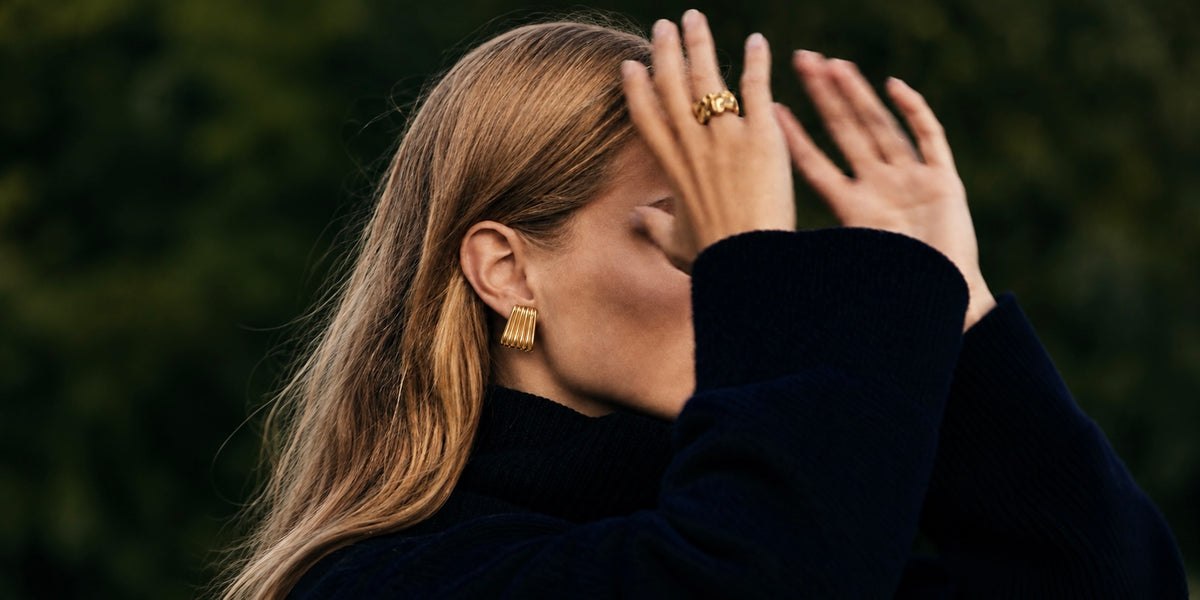 A woman with long blonde hair wearing a black turtleneck sweater and gold geometric earrings, posing outdoors with her hands covering part of her face against a blurred green background.